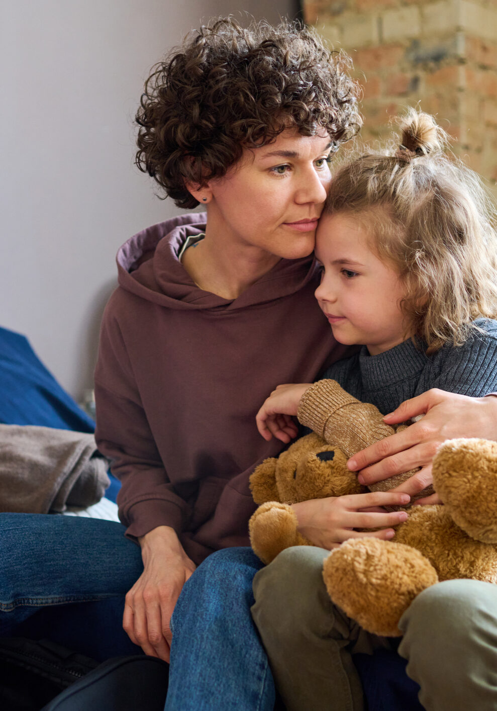 Young woman giving hug to her cute little son with brown soft teddybear while both sitting on sleeping place prepared for refugees